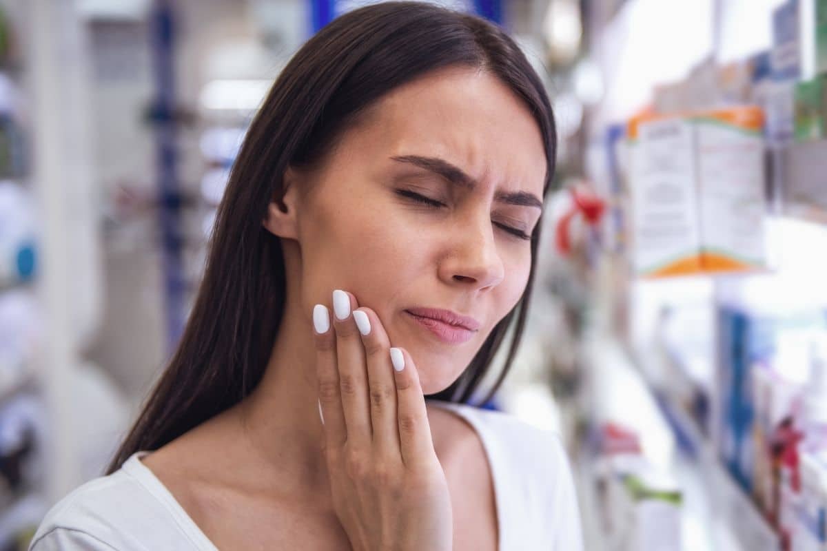 Woman with tooth pain holding her cheek in a pharmacy setting.