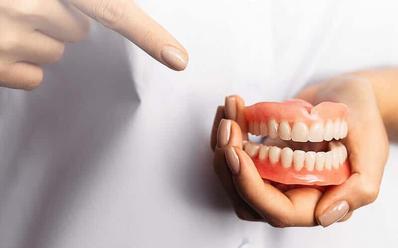 Hands holding a model of full dentures showing upper and lower teeth with red gum tissue.